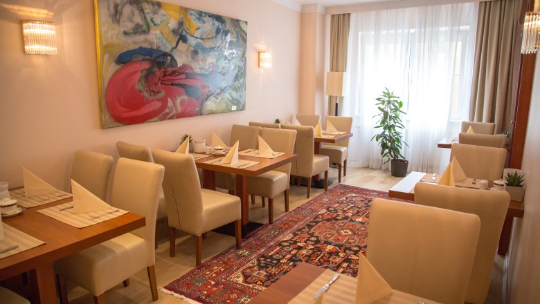 Frühstücksraum, © Hotel Admiral am Kurpark Ein eleganter Frühstücksraum mit gedeckten Tischen, beigen Stühlen, einem bunten Teppich und einem großen abstrakten Gemälde an der Wand.