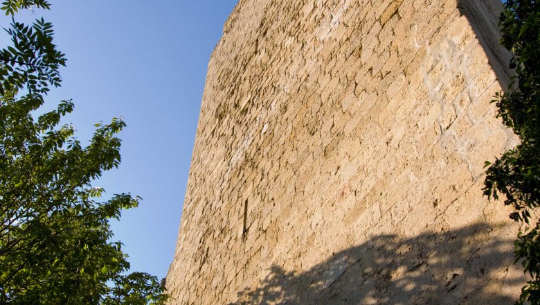 Nahaufnahme eines hohen, alten Burgturms aus Stein, umgeben von B&auml;umen, unter blauem Himmel.