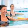 Two women are doing aqua aerobics in a pool with yellow swimming noodles.