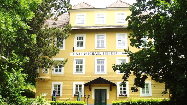 Front view of the Carl Michael Ziehrer House, a yellow building with several windows and a green entrance, surrounded by trees.