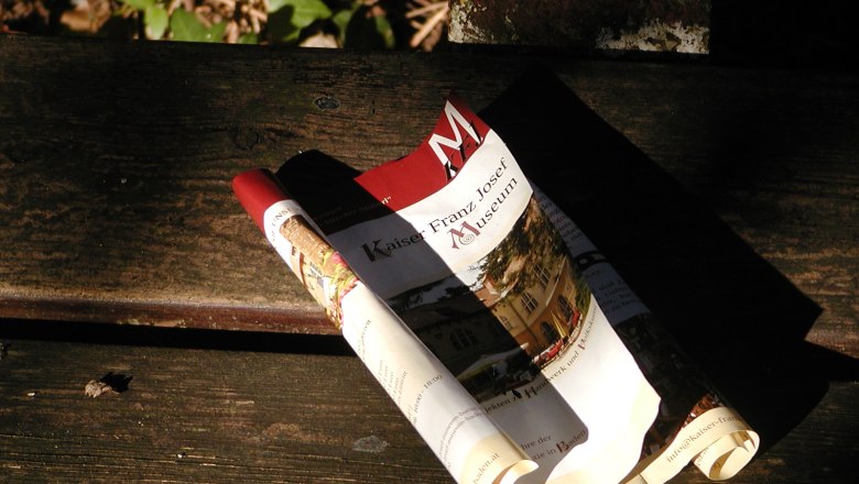 Zusammengerolltes Prospekt des Kaiser-Franz-Josef-Museums auf einer Holzbank.