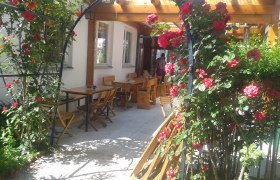 A rose arch with red roses leads to an outdoor area with wooden tables and chairs.
