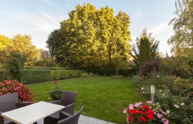 Ein gepflegter Garten mit Rasen, Blumenbeeten und Gartenm&ouml;beln auf einer Terrasse.